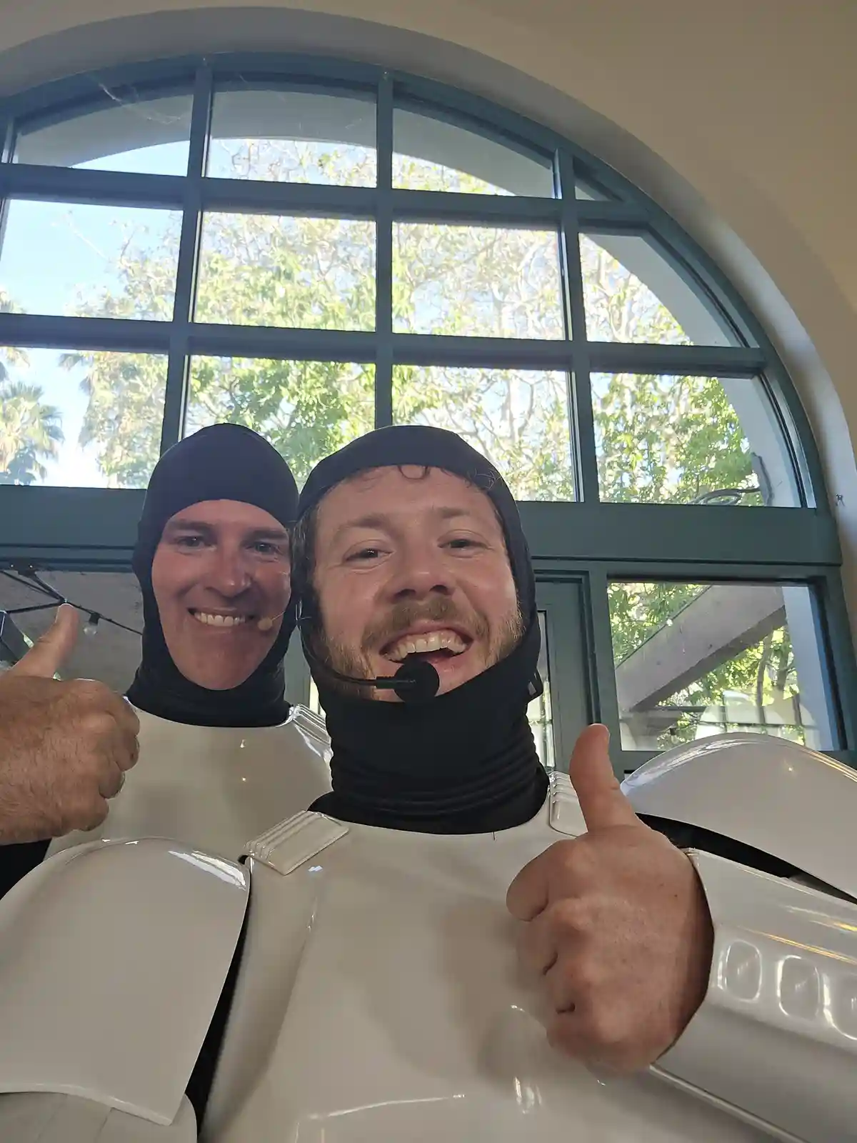 Two men in white robot costumes with black balaclavas and headsets are smiling and giving thumbs up to the camera. Behind…