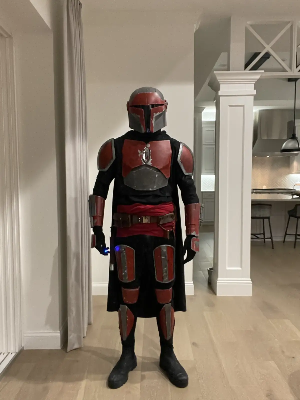 A person in a detailed Mandalorian armor costume, featuring red and black plating with a helmet, stands in a modern home…