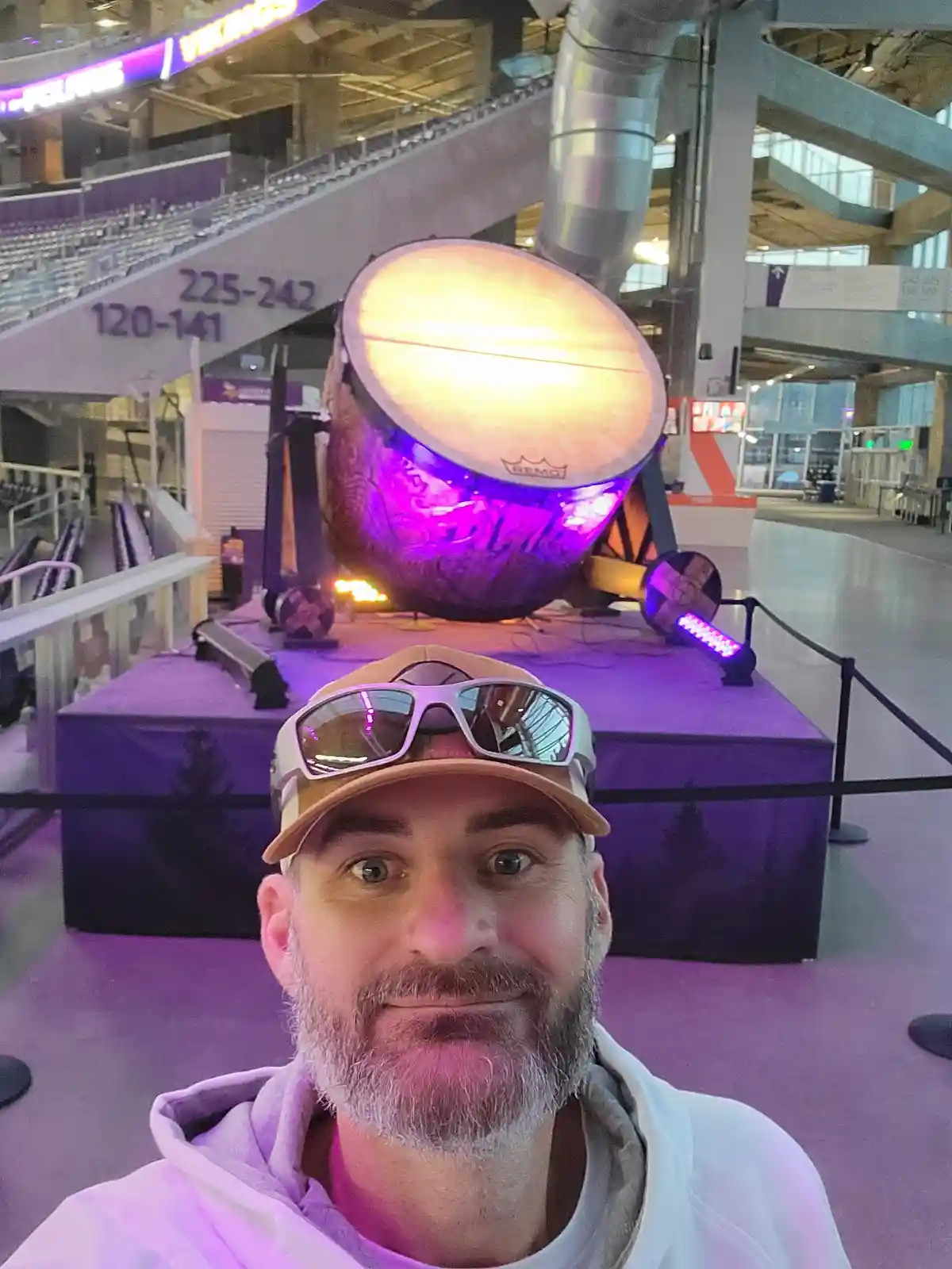 A man with a beard and sunglasses on his cap takes a selfie in front of a large, illuminated purple drum at a stadium. The…