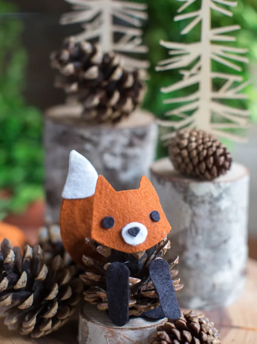 A handmade felt fox face with white-tipped ears and black eyes is glued onto a pinecone, sitting on a birch wood slice…