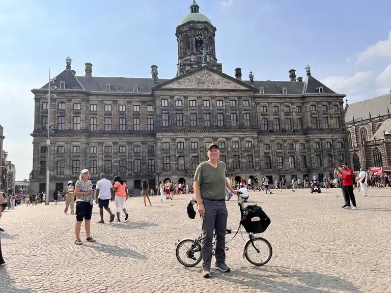 A man in a green shirt and cap stands smiling in front of a large historic building with a dome and clock tower, holding a…