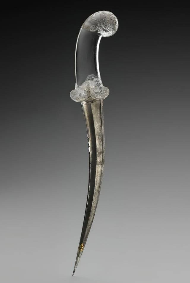 a clear glass dagger with an ornate, curved hilt and decorative floral carvings, set against a gradient gray background. The…