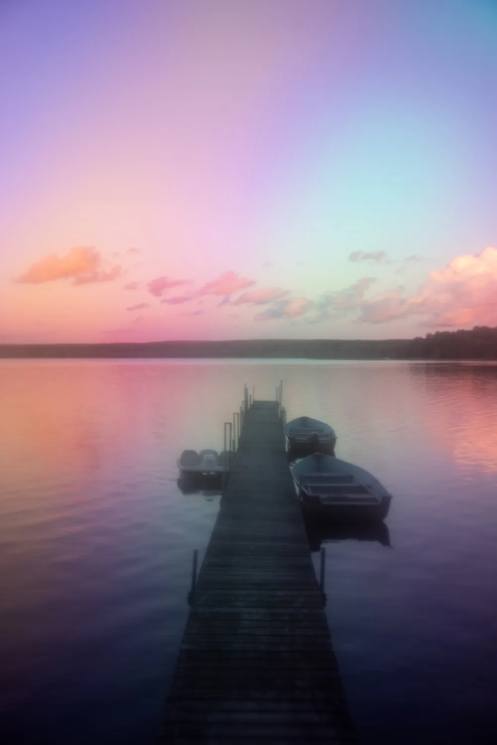 A serene, long-exposure photograph of a wooden dock extending into a calm lake at sunset, with boats moored alongside. The…