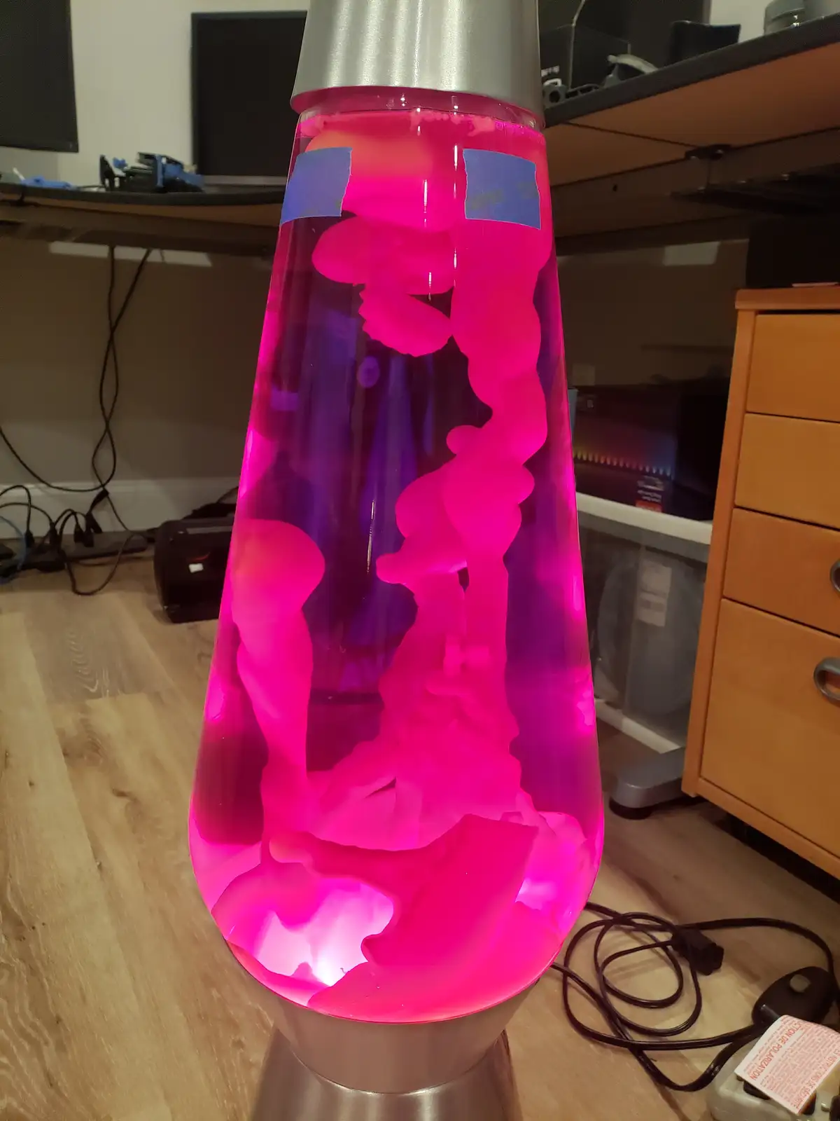 A pink lava lamp with a silver base and top sits on a wooden floor in a room with desks and electronics. Two small blue tape…
