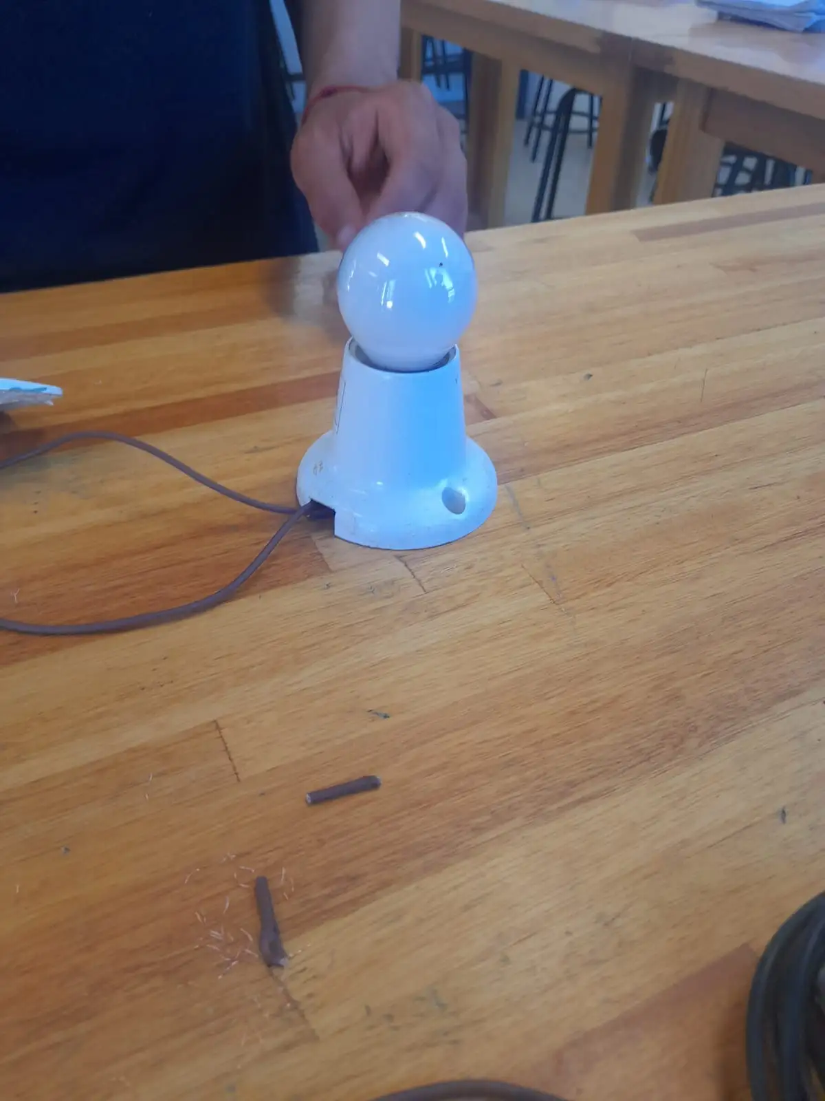 A person's hand hovers over a white light bulb screwed into a white socket on a wooden table, with a brown wire extending…