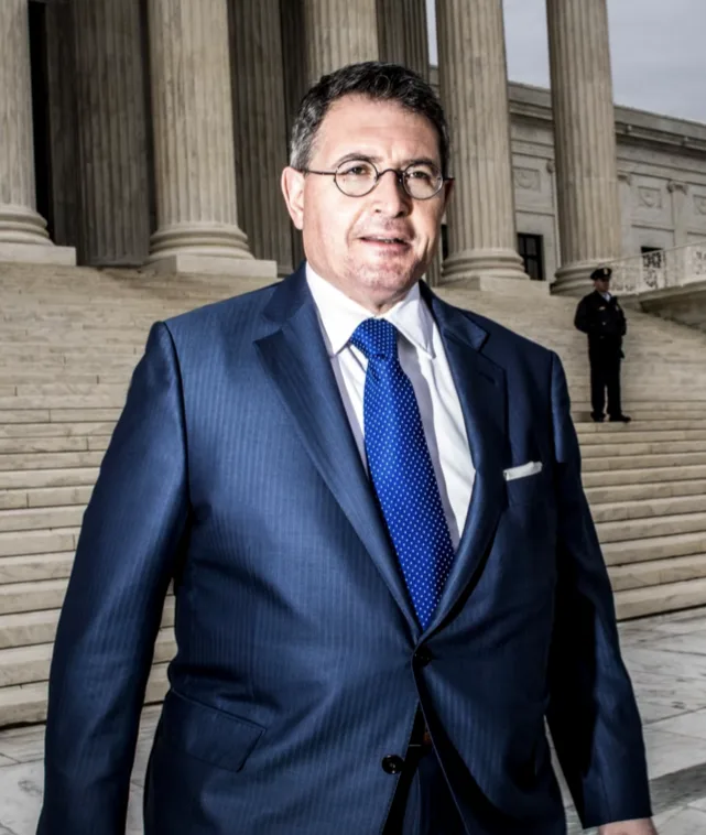 A man in a blue suit and glasses stands confidently in front of the Supreme Court building, with a guard visible in the…