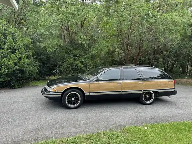 A black and wood-paneled station wagon, likely a Ford Taurus wagon, is parked on an asphalt driveway surrounded by green…