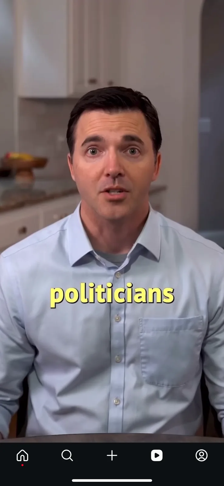 A man in a light blue button-down shirt sits in what appears to be a kitchen, looking directly at the camera with a…