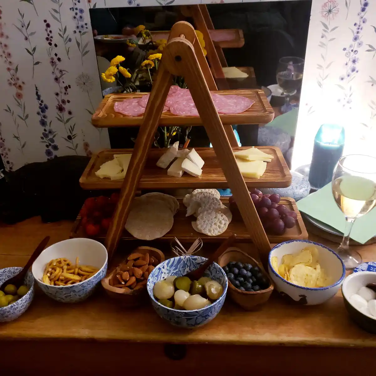 A wooden three-tiered serving stand holds sliced salami, various cheeses, crackers, grapes, and tomatoes. Surrounding it on…