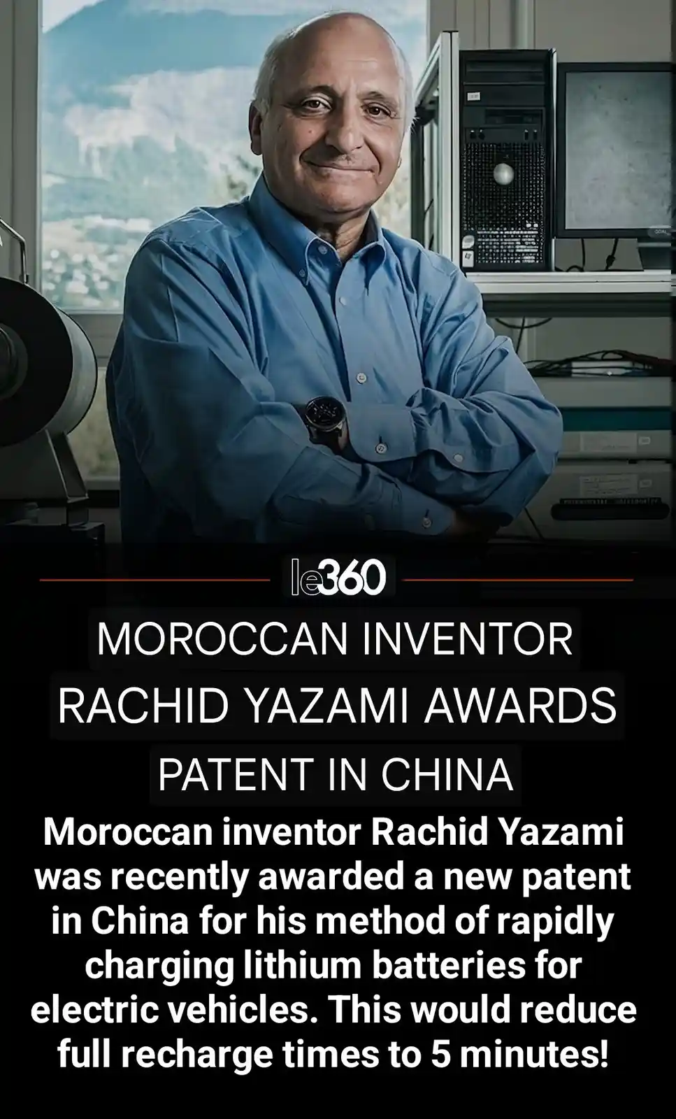 A smiling man in a blue shirt stands with arms crossed in a lab setting with computer equipment and a mountain view…