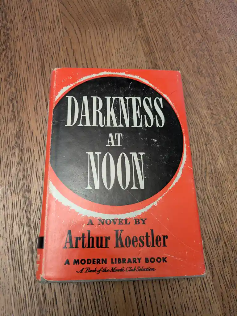 A worn, red-covered paperback book titled 'DARKNESS AT NOON' by Arthur Koestler rests on a wooden surface. The cover…