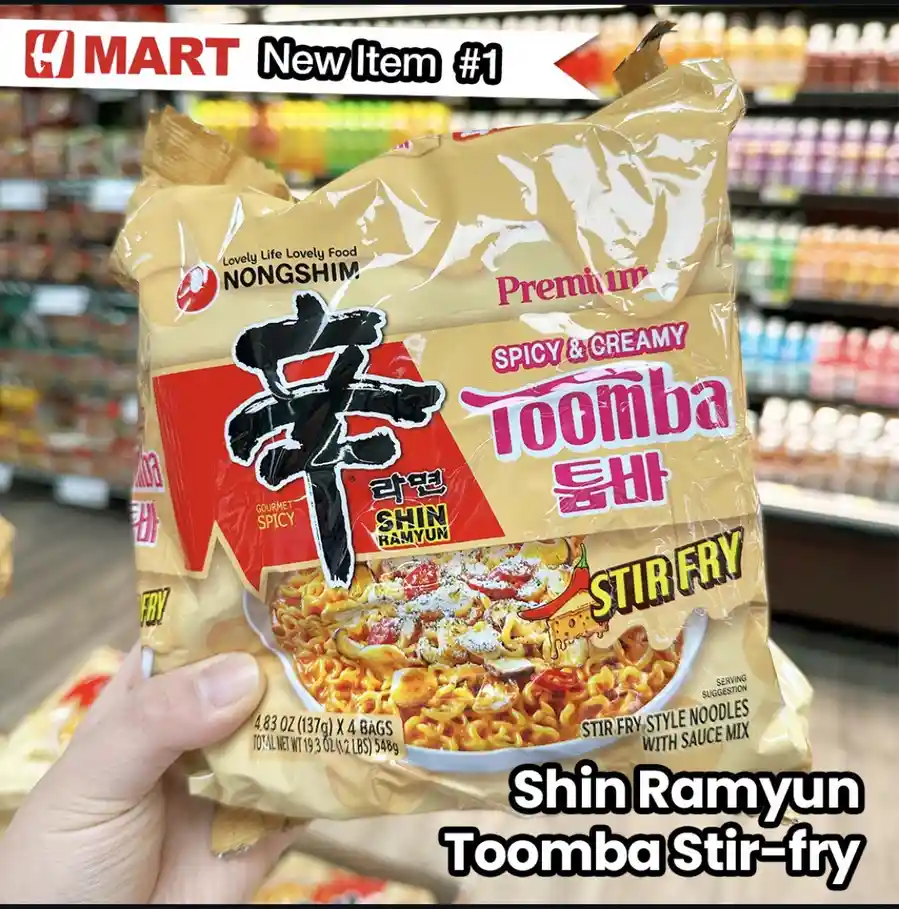 A hand holds a large package of Nongshim Shin Ramyun Toomba Stir-fry noodles in a grocery store aisle. The packaging is gold…