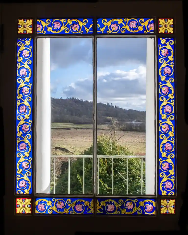 A view through an ornate, colorful stained-glass window with blue, gold, and pink floral patterns, looking out onto a cloudy…