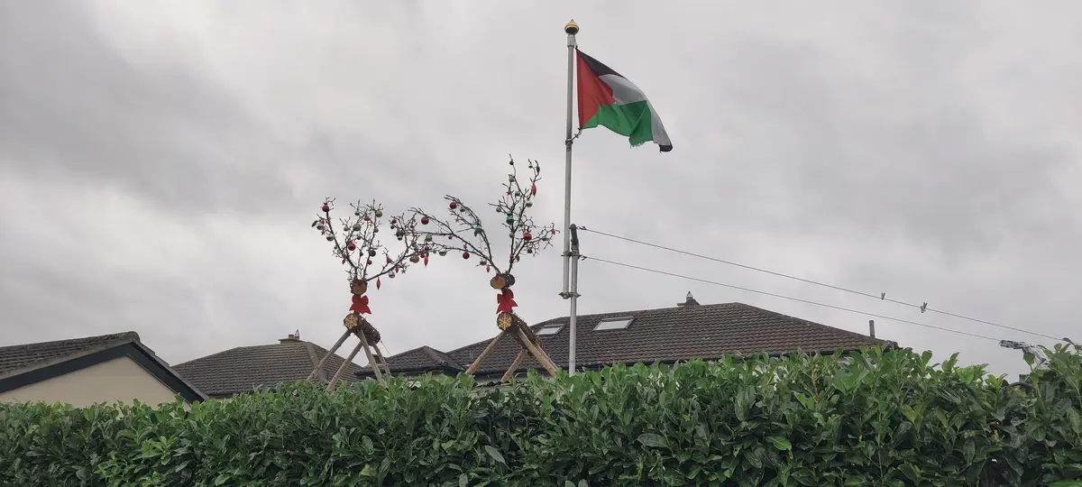 A Palestinian flag flies from a tall pole next to two decorative, bare-branched reindeer figures adorned with Christmas…
