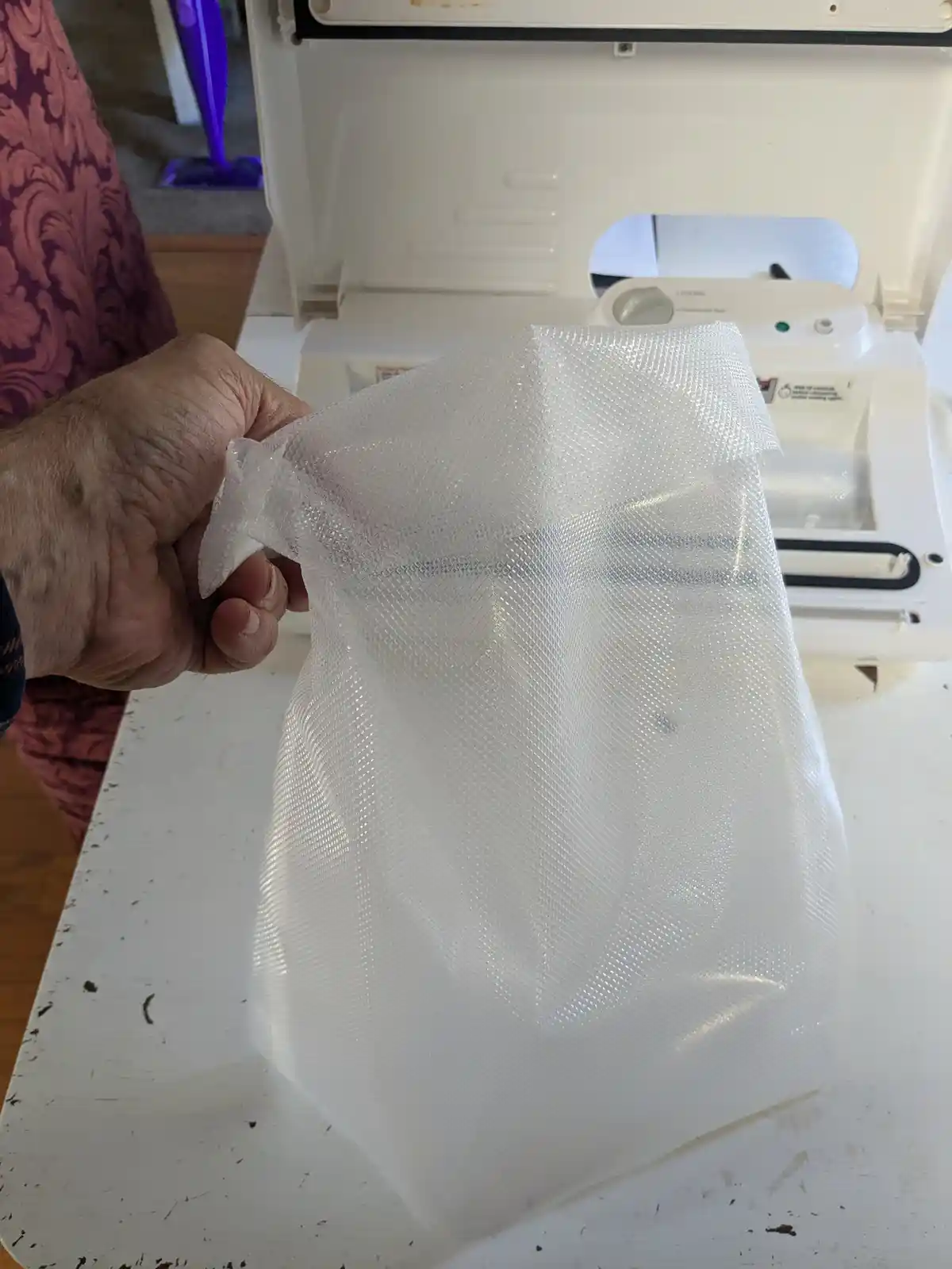 A person's hand holds a white, textured vacuum bag in front of a white vacuum cleaner with its top open. The bag appears…