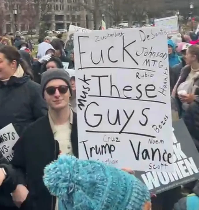 A person at a protest holds a large white sign with handwritten text that reads 'Fuck These Guys' in large letters, with…