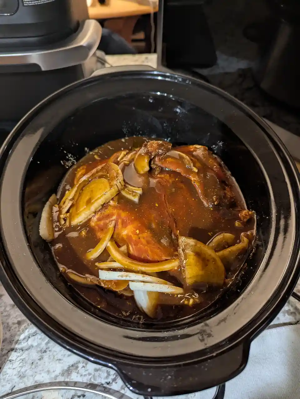 A black slow cooker contains a large piece of meat, likely pork or beef, submerged in a dark brown sauce with sliced onions…