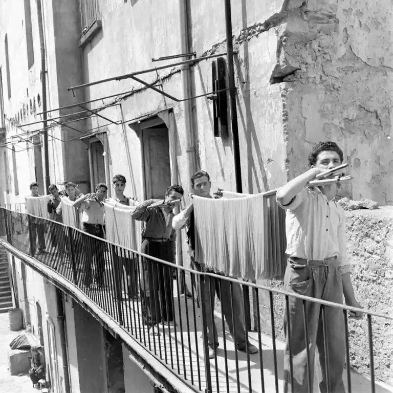 a black and white historical photo shows a line of men standing on a narrow outdoor balcony, each holding a long, flat…