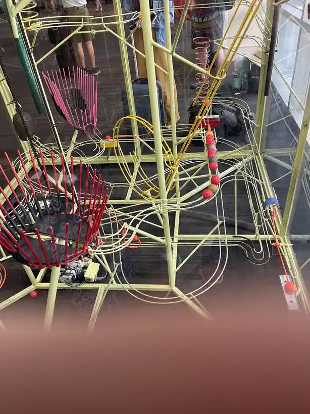 A complex, colorful mechanical sculpture with red and pink spiky elements, yellow wires, and red balls rolling down tracks…