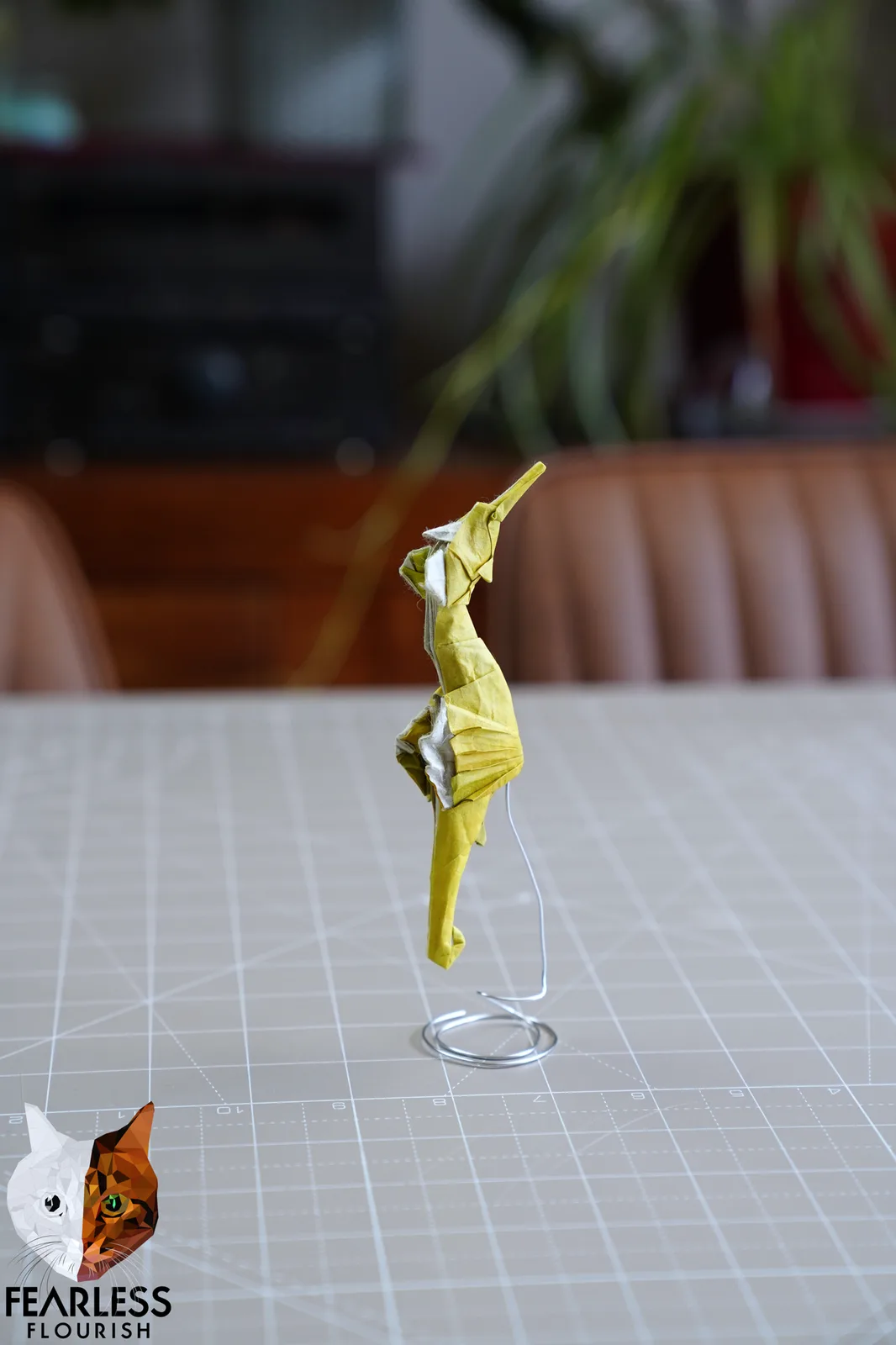A yellow origami seahorse stands upright on a silver wire stand placed on a gray gridded cutting mat. The background is…