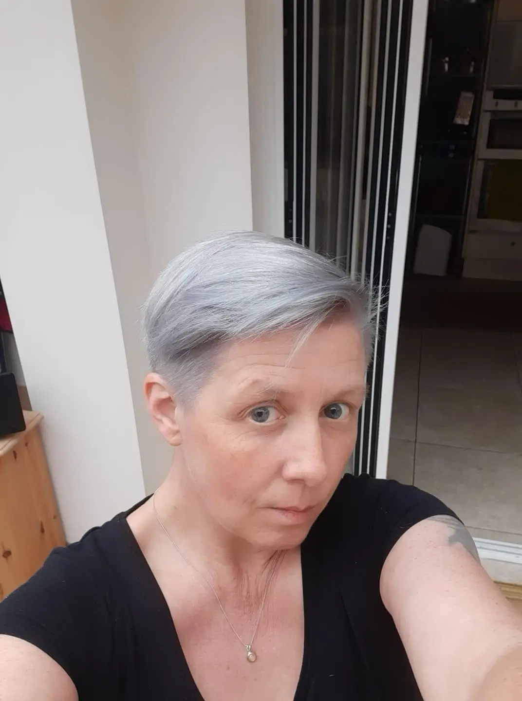 A selfie of a woman with short, silver-grey hair, wearing a black top and a pearl necklace, looking directly at the camera…