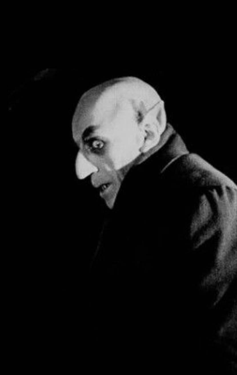 A black-and-white image shows a bald man with pointed ears, a long nose, and a menacing expression, looking over his…
