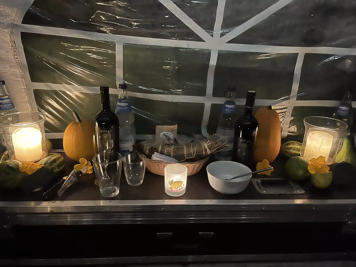 A dimly lit, rustic bar setup inside a tent with plastic windows, featuring pumpkins, gourds, wine bottles, water bottles…