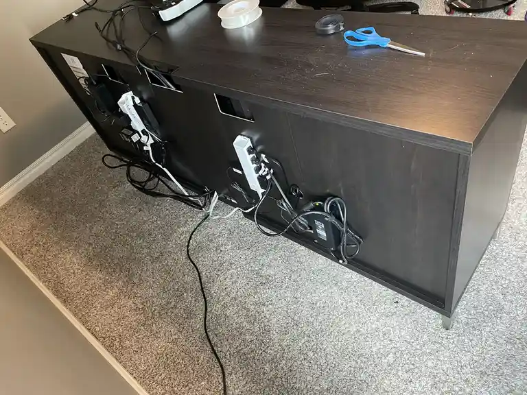 A dark wooden media console has numerous tangled power strips and cables plugged into its back, spilling onto the carpeted…