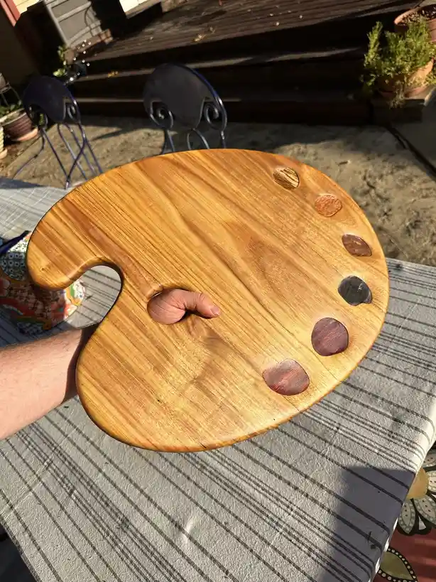 A person holds up a wooden artist's palette with a thumb inserted into its thumb hole, showing off the palette's natural…