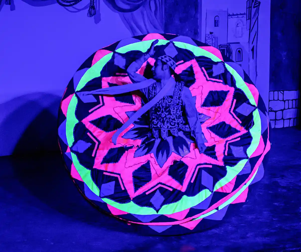 A person in traditional attire is performing a dance inside a large, circular, neon-pink and green patterned costume that…