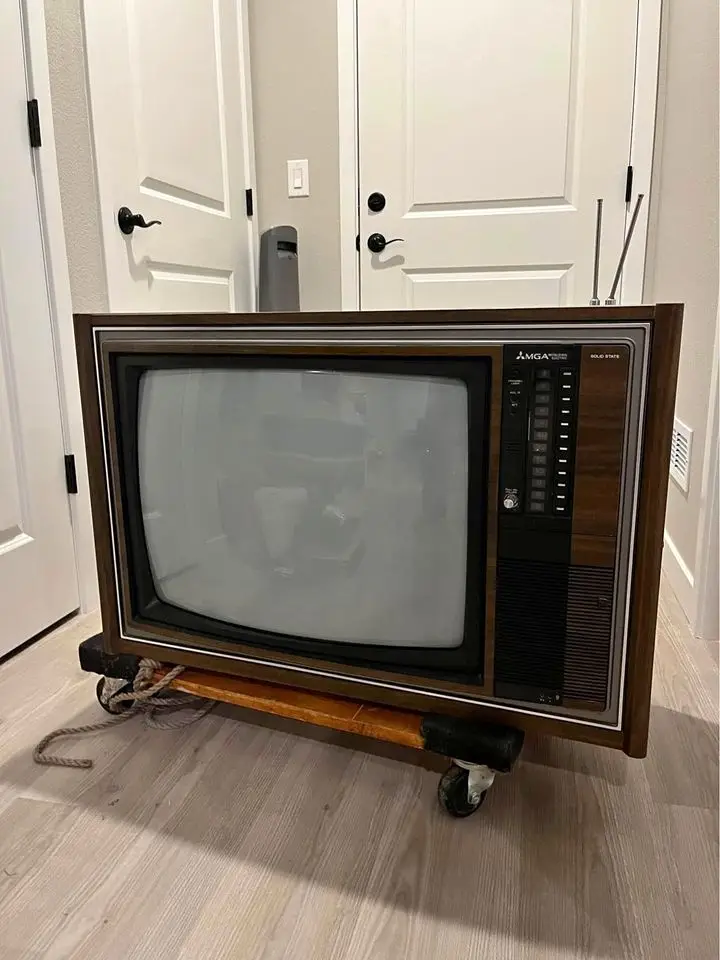A vintage wooden-cased AMGA television with rabbit ears sits on a wheeled dolly in a modern home, with a rope tied to the…