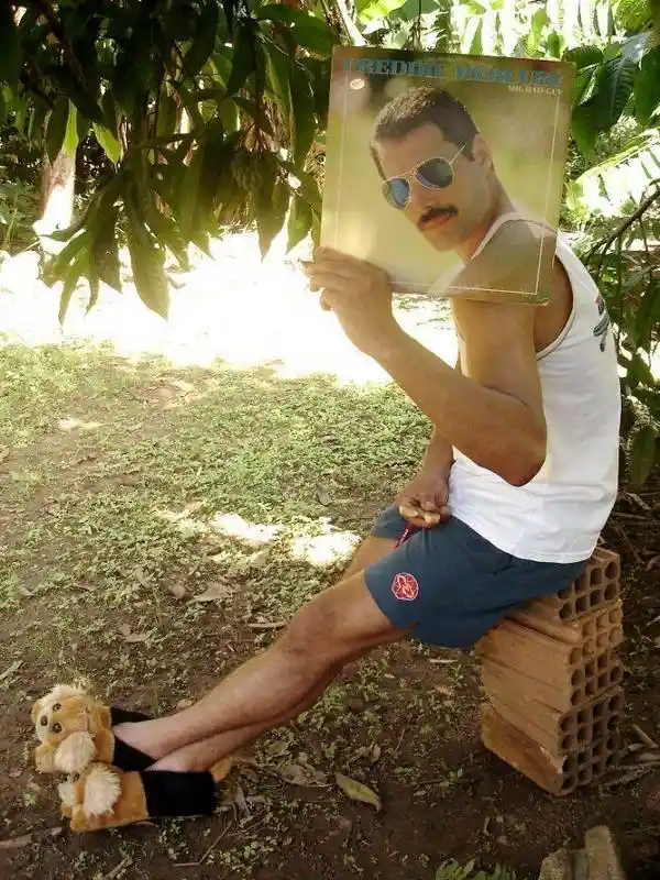 A man sits outdoors on a stack of bricks wearing a white tank top and blue shorts, holding up a Freddie Mercury album cover…