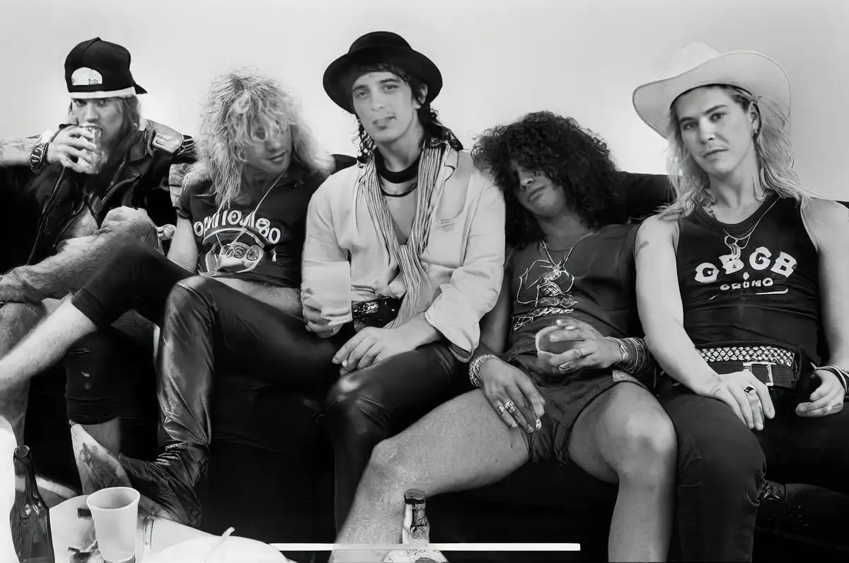 A black-and-white photo of five men in 1980s glam metal attire sitting on a couch, with one man in the center looking…
