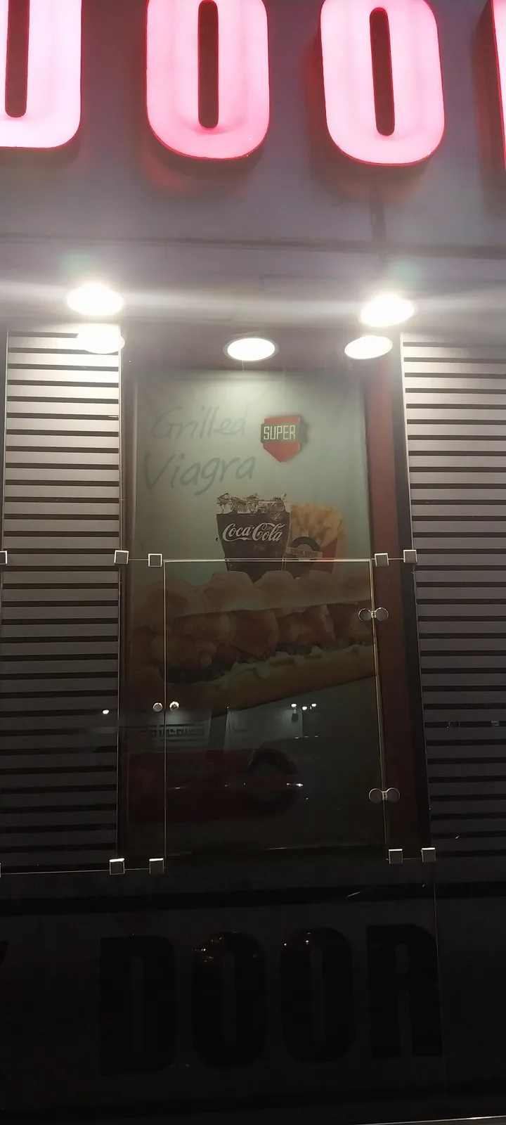 A photo of a restaurant's glass door with a poster advertising a sandwich labeled 'Grilled Viagra' next to a Coca-Cola and…