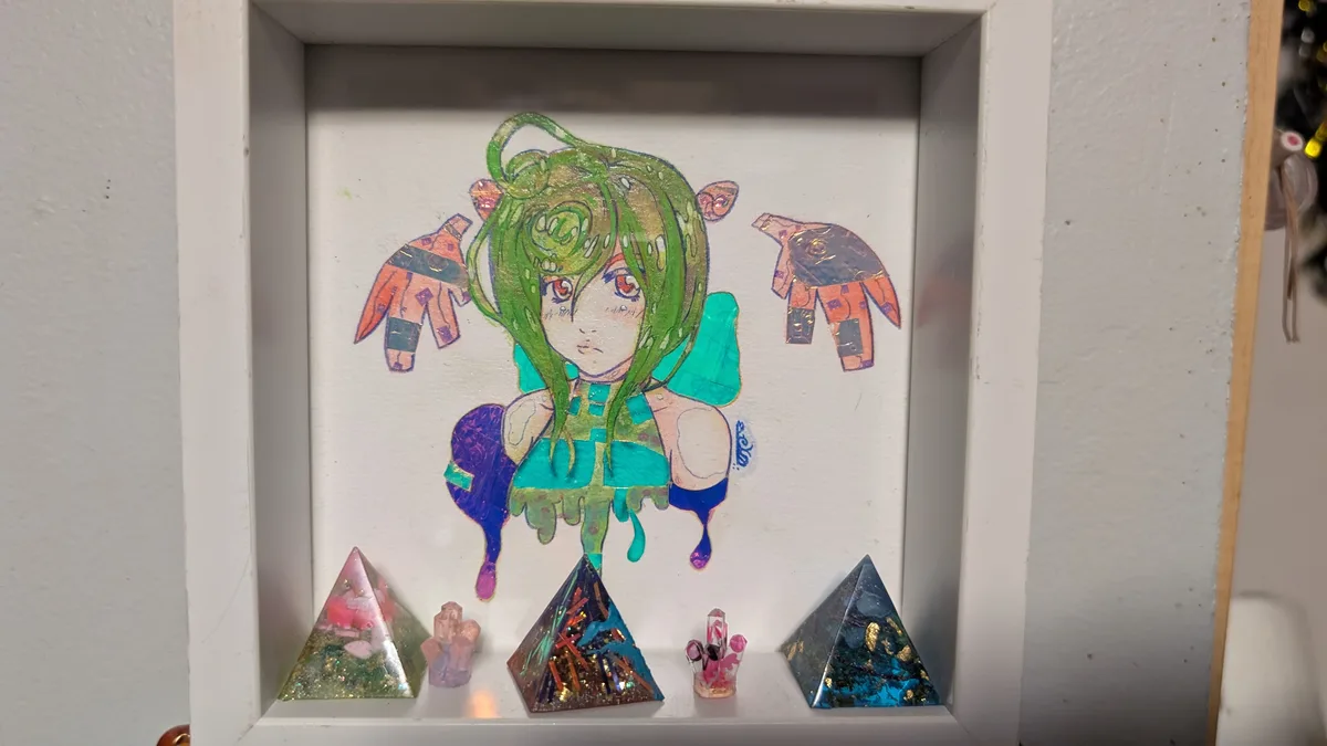 A white shadow box frame contains a colorful anime-style drawing of a girl with green hair and red eyes, wearing a teal…