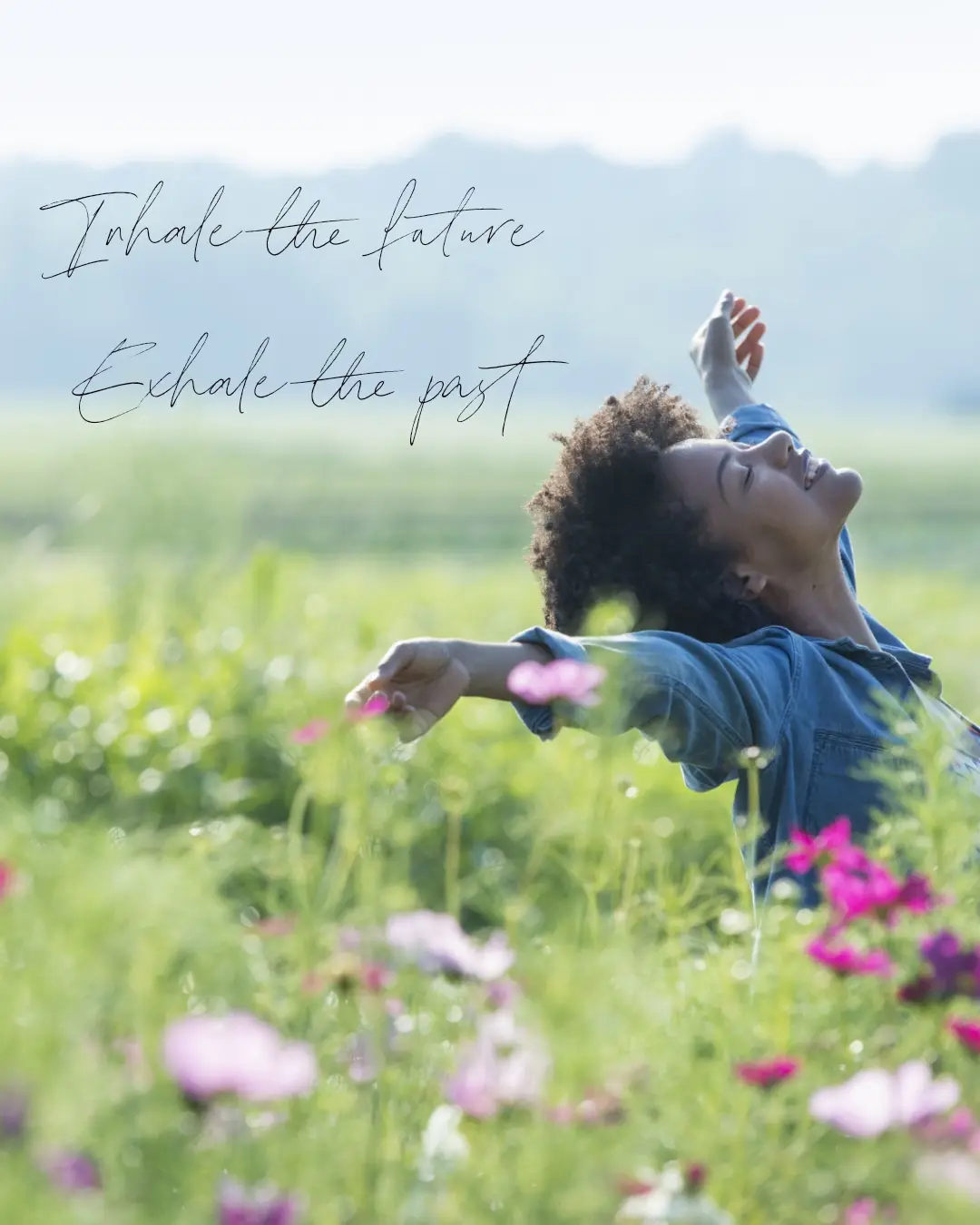 A person with curly hair, wearing a denim shirt, stands in a sunlit field of green grass and pink cosmos flowers, arms…