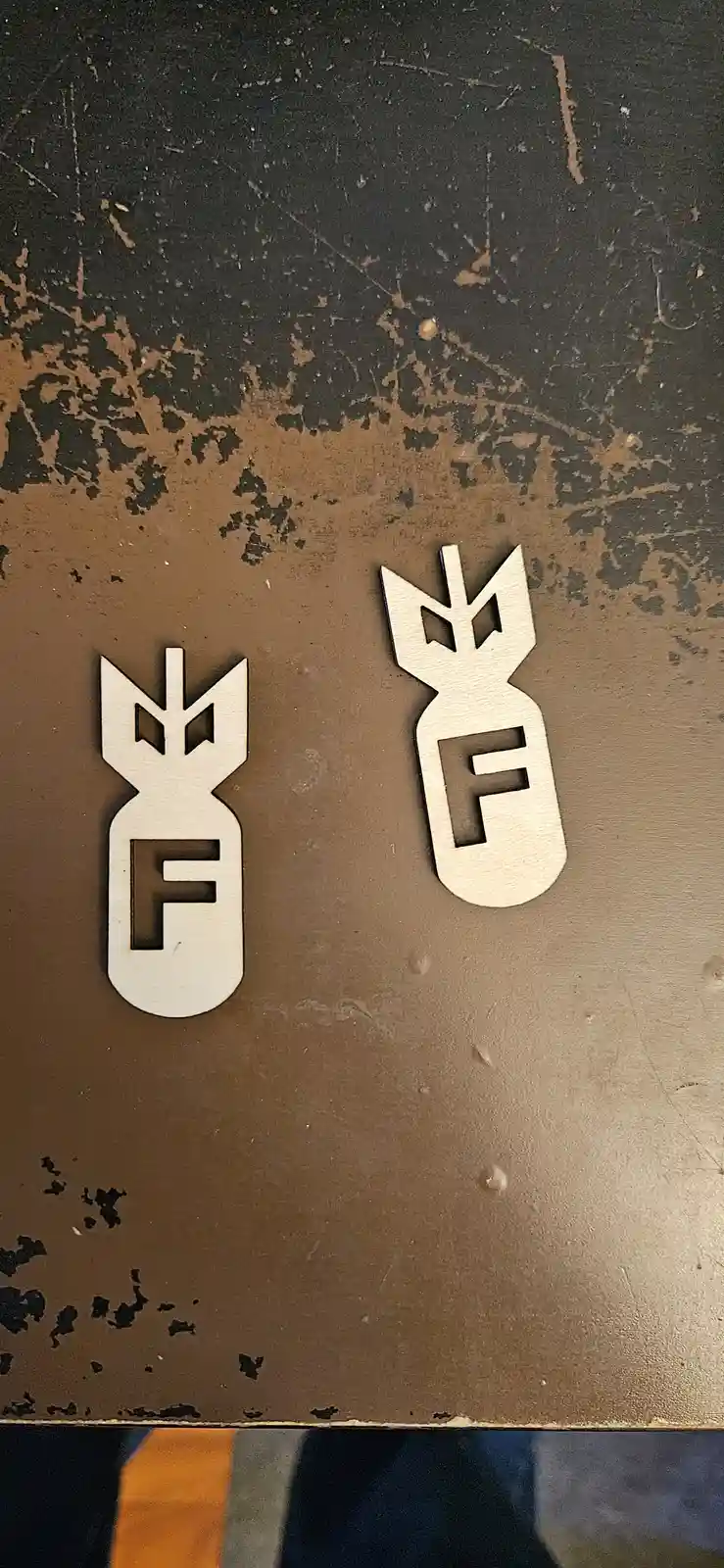 Two light-colored, laser-cut bomb-shaped objects sit on a worn, dark brown surface. Each bomb has the letter 'F' cut out…