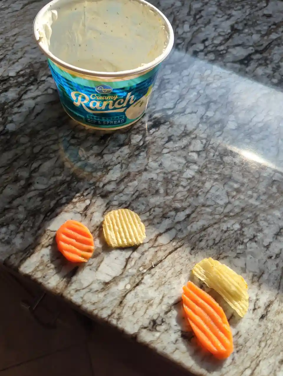 An empty Kroger Creamy Ranch dip container sits on a marble countertop next to several waffle-cut potato chips and carrot…