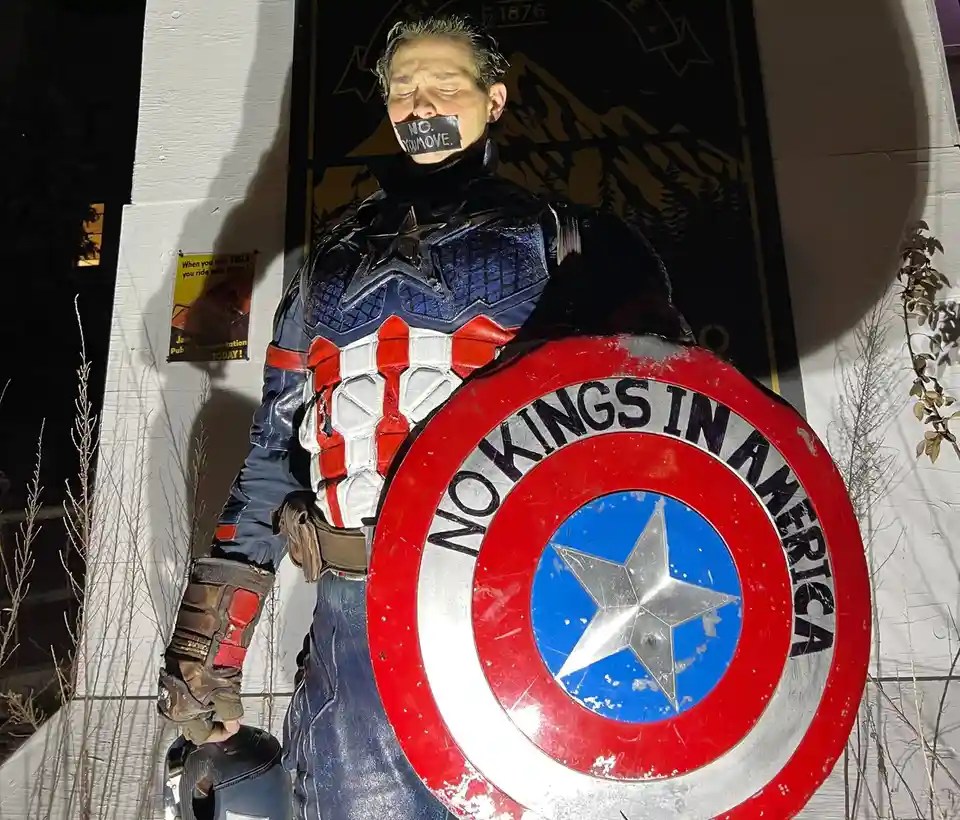 A person in a Captain America costume stands with eyes closed and mouth covered by tape reading 'NO. YOU MOVE.' Their shield…