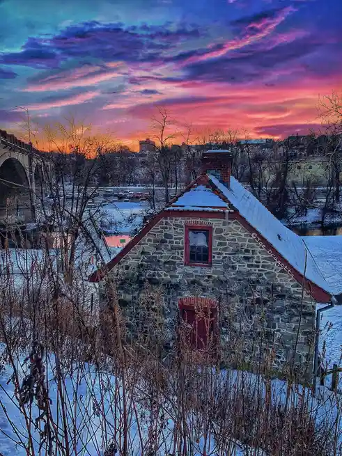 A small stone house with red trim and a snow-covered roof sits in a snowy landscape under a vibrant, colorful sunset sky…
