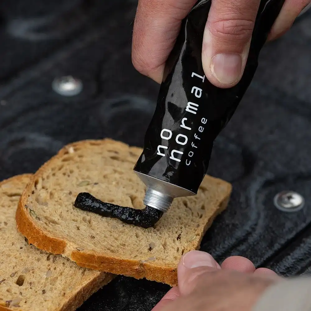 A hand squeezes a black tube labeled 'normal coffee' to dispense dark coffee paste onto a slice of whole grain bread…