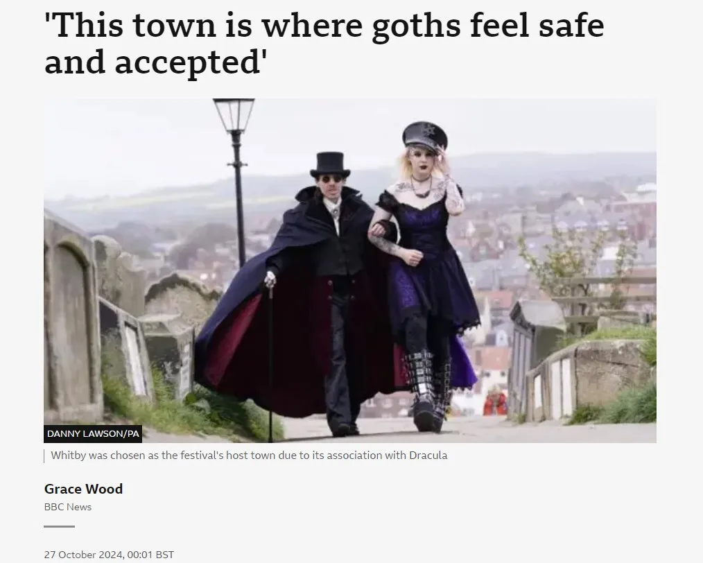 Two people dressed in gothic fashion—a man in a top hat and cape, and a woman in a purple dress and cap—walk on a path…