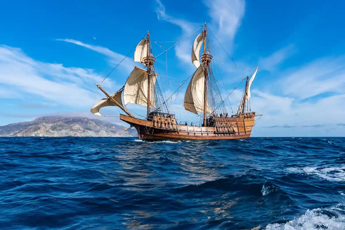 A wooden sailing ship with multiple masts and white sails glides across deep blue ocean waters under a bright blue sky with…