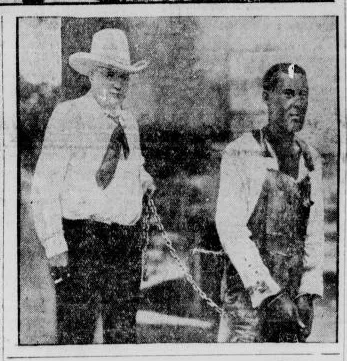 A grainy, black-and-white historical photo shows a man in a cowboy hat holding a chain that is attached to another man…