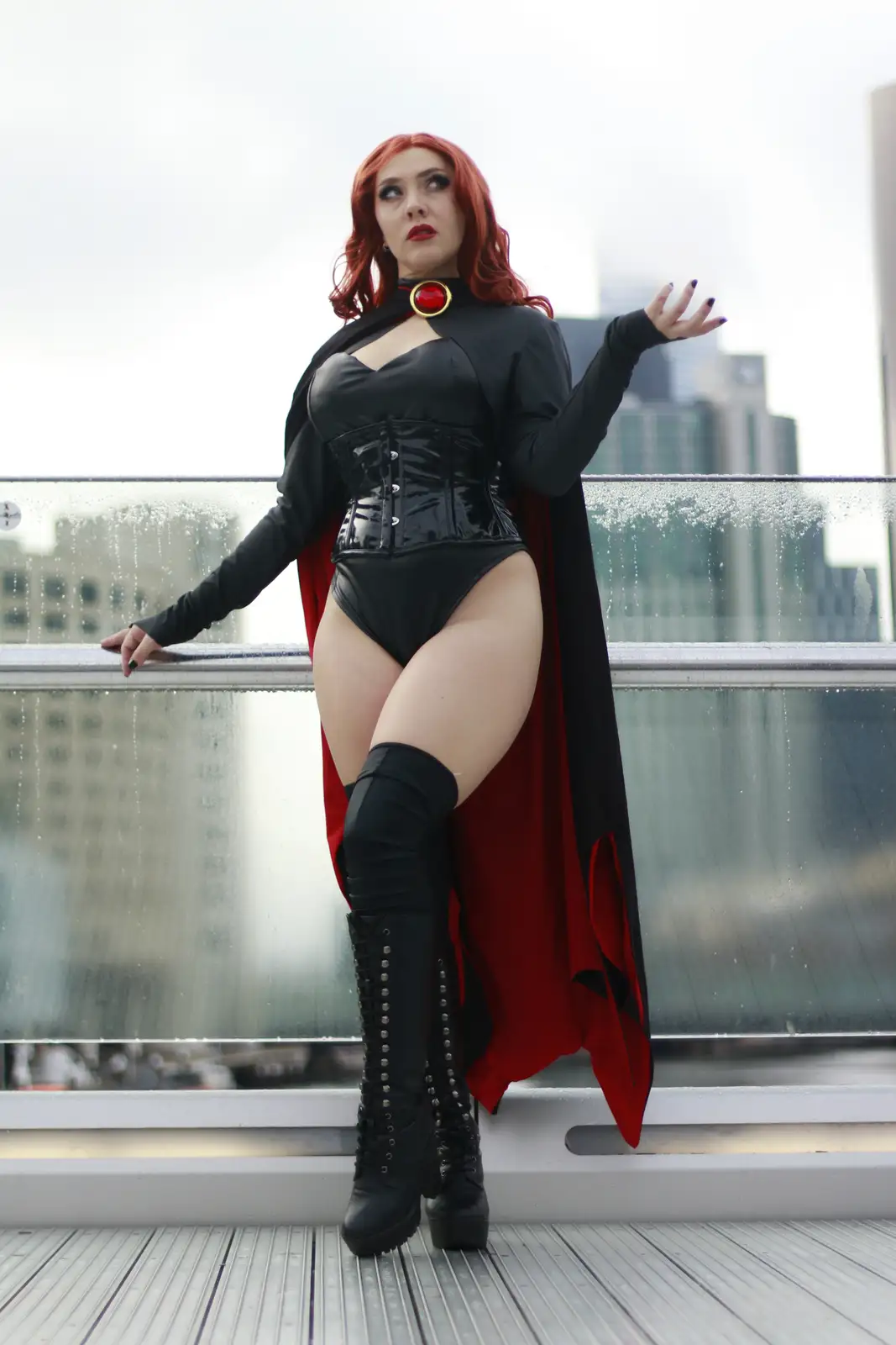 A woman in a black and red superhero costume with a cape and a red gemmed collar poses on a wet glass balcony overlooking a…
