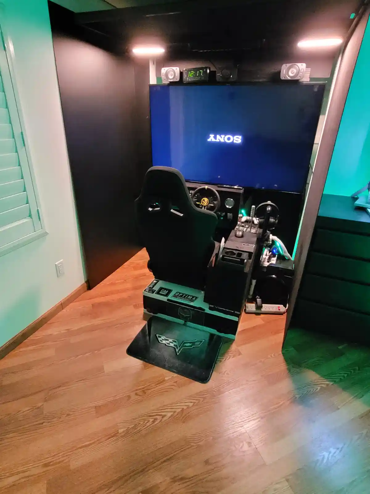 A high-end racing simulator setup with a black racing seat, steering wheel, and pedals, positioned in front of a large TV…