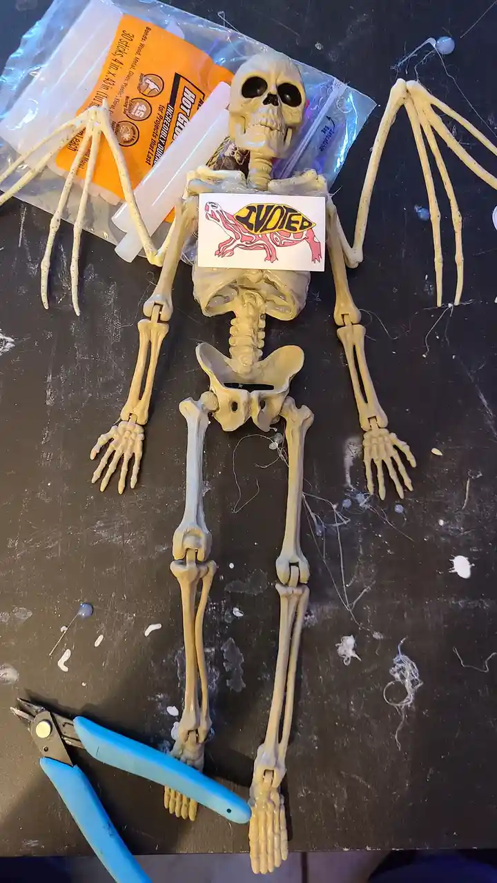 A plastic skeleton model lies on a cluttered workbench surrounded by crafting supplies like hot glue sticks, pliers, and…