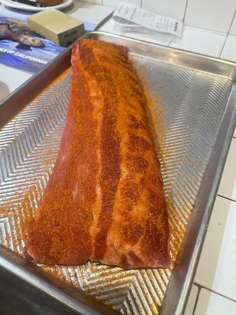 A large raw slab of ribs, heavily coated in orange-red seasoning, sits on a metal baking tray with a zigzag pattern. The…