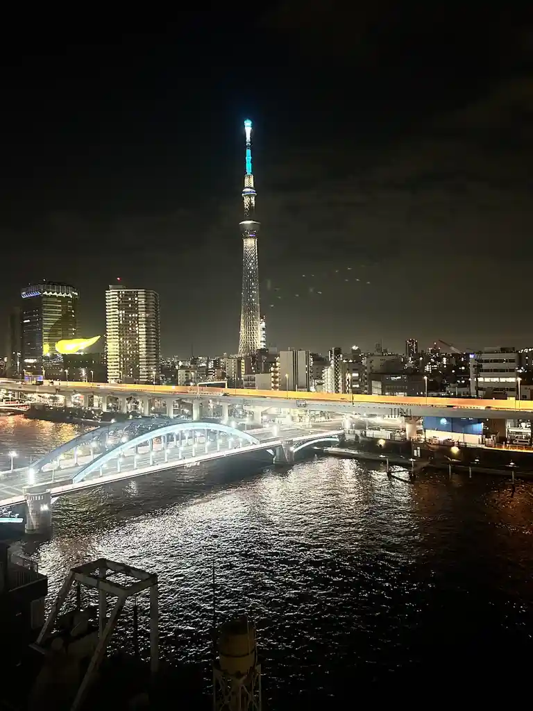 A nighttime cityscape photo featuring the illuminated Tokyo Skytree tower, a river with a lit blue arched bridge, and…