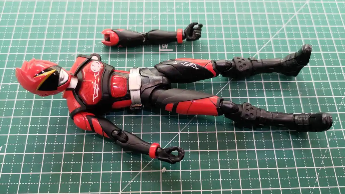 A red and black Kamen Rider action figure lies flat on a green cutting mat, with its left arm detached and placed beside its…